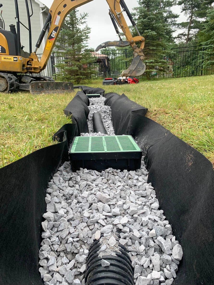 Drainage installation in a backyard: trench lined with filter fabric, gravel, drain pipe and a catch basin, with a mini excavator behind
